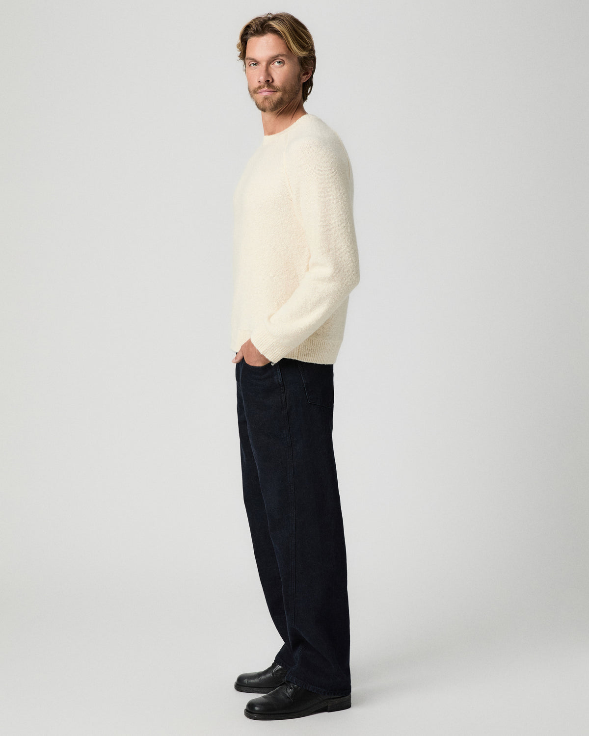 Side profile of model with one hand in his pocket, wearing the Hightower Crewneck Sweater in Marfil with dark relaxed jeans and black boots