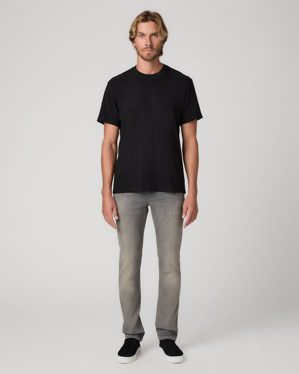 A man modeling a black short-sleeve crewneck t-shirt, light gray slim-fit jeans, and black slip-on shoes against a plain light gray studio background.