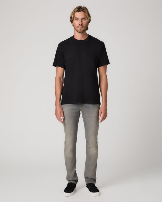 A man modeling a black short-sleeve crewneck t-shirt, light gray slim-fit jeans, and black slip-on shoes against a plain light gray studio background.