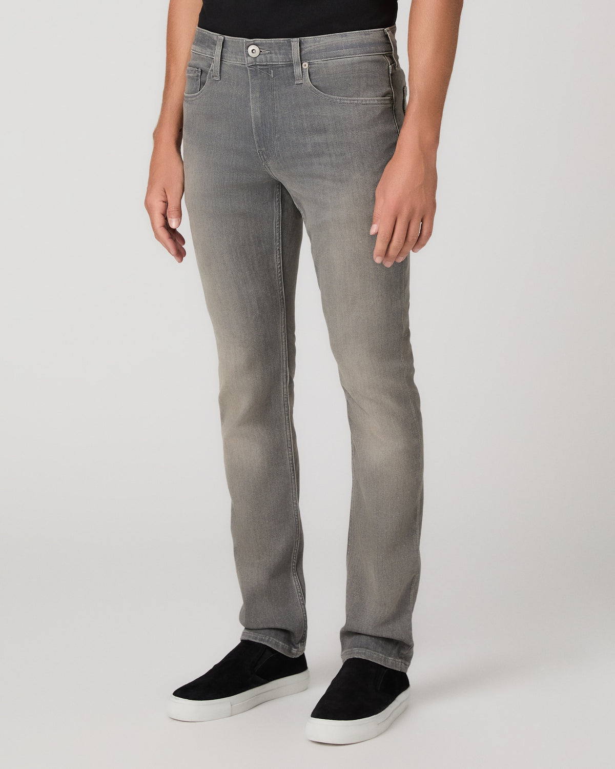 A man modeling a black short-sleeve crewneck t-shirt, light gray slim-fit jeans, and black slip-on shoes against a plain light gray studio background.