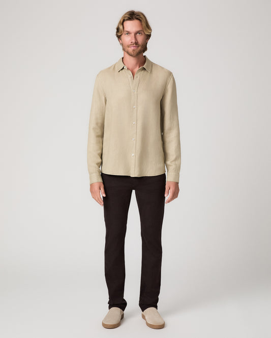 A man modeling a light beige linen button-down shirt with dark brown trousers and beige loafers, standing against a neutral light gray studio background.