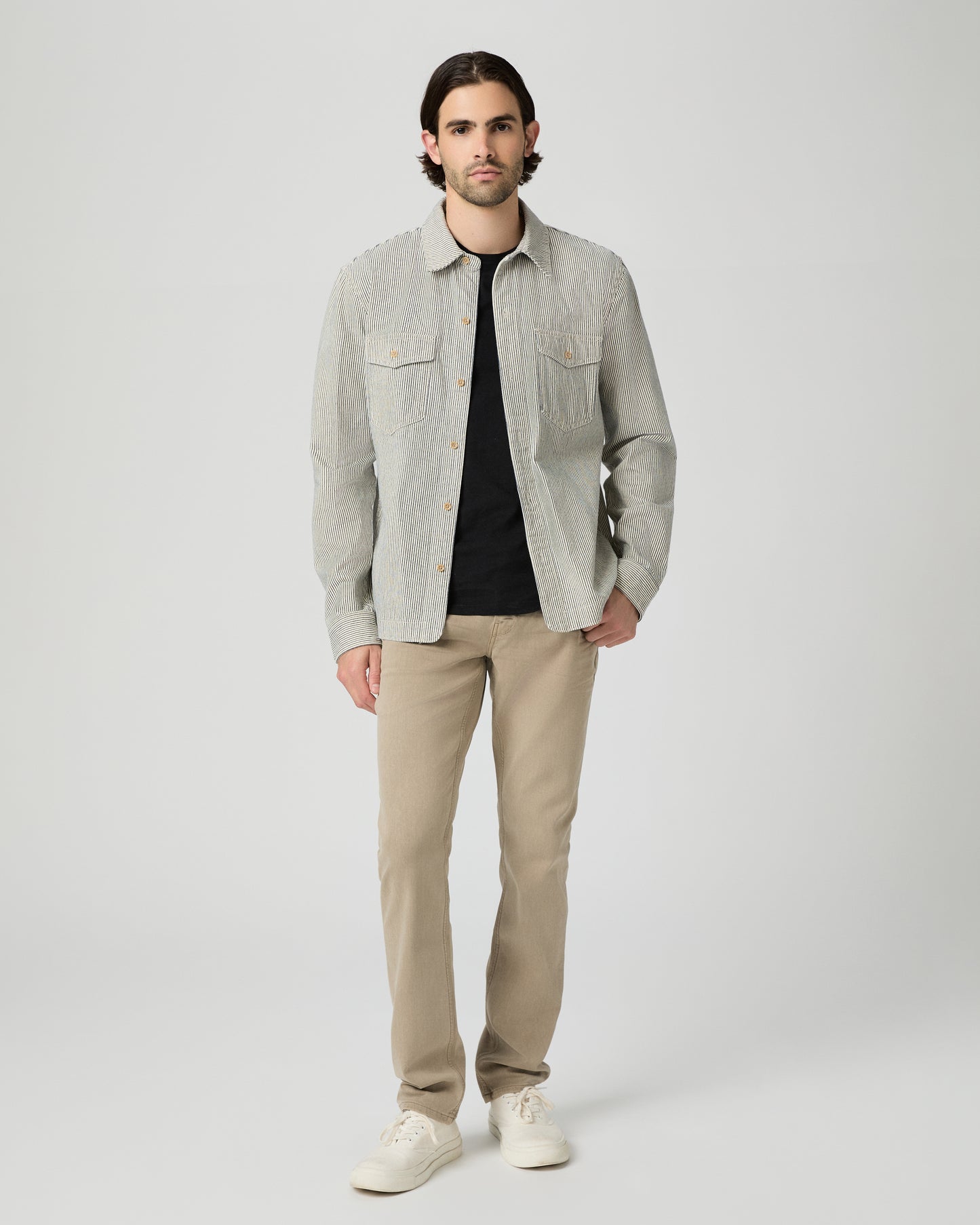 Man wearing a light gray shirt over a black t-shirt, beige pants, and white shoes on a white background
