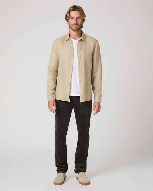 A man modeling a light beige linen button-up shirt over a white t-shirt, paired with black jeans and beige slip-on shoes, against a neutral light gray studio background.