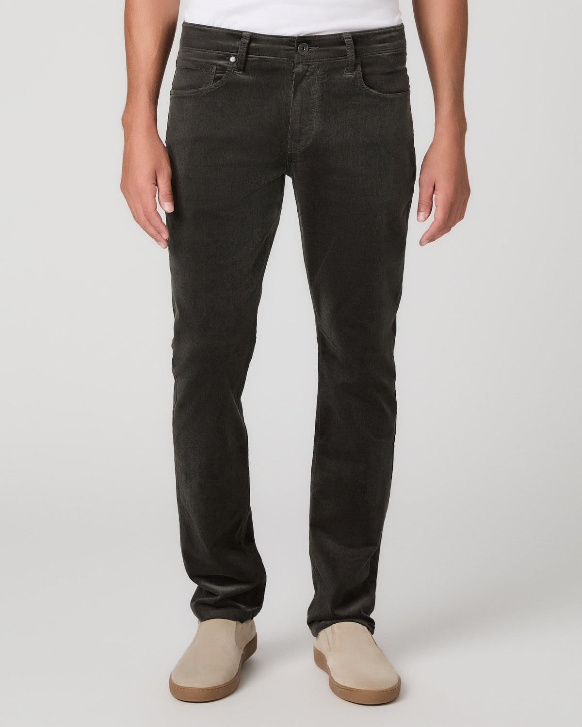 A man modeling a white t-shirt, paired with black jeans and beige slip-on shoes, against a neutral light gray studio background.