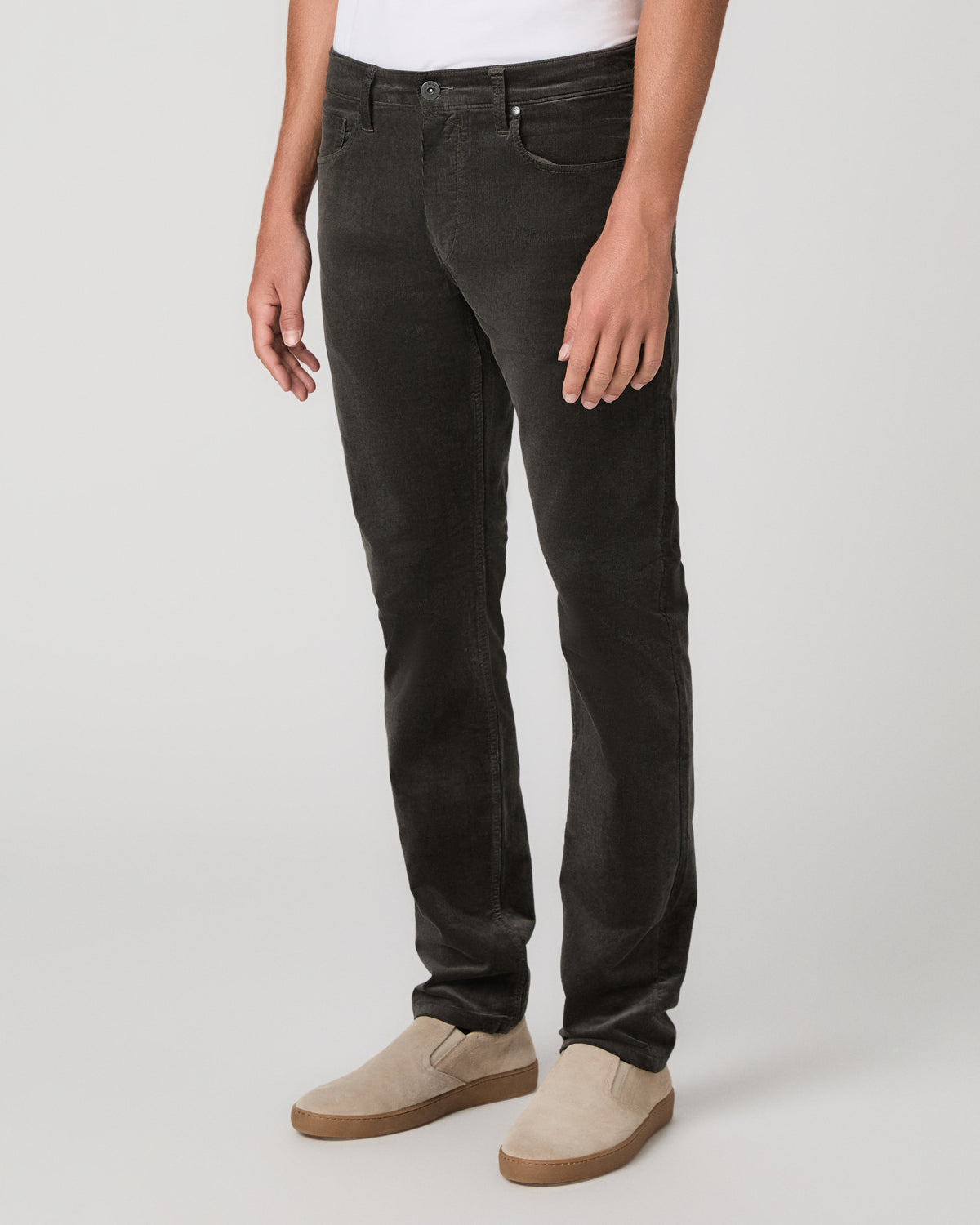 A man modeling a white t-shirt, paired with black jeans and beige slip-on shoes, against a neutral light gray studio background.