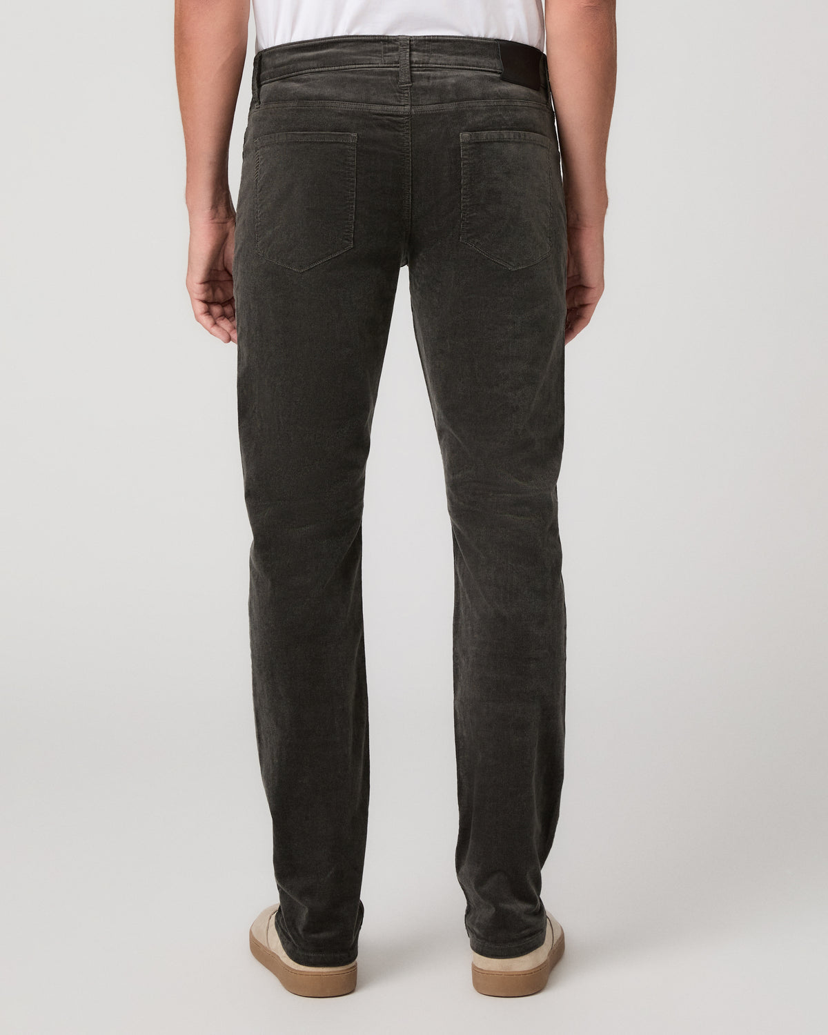 A man modeling a white t-shirt, paired with black jeans and beige slip-on shoes, against a neutral light gray studio background.