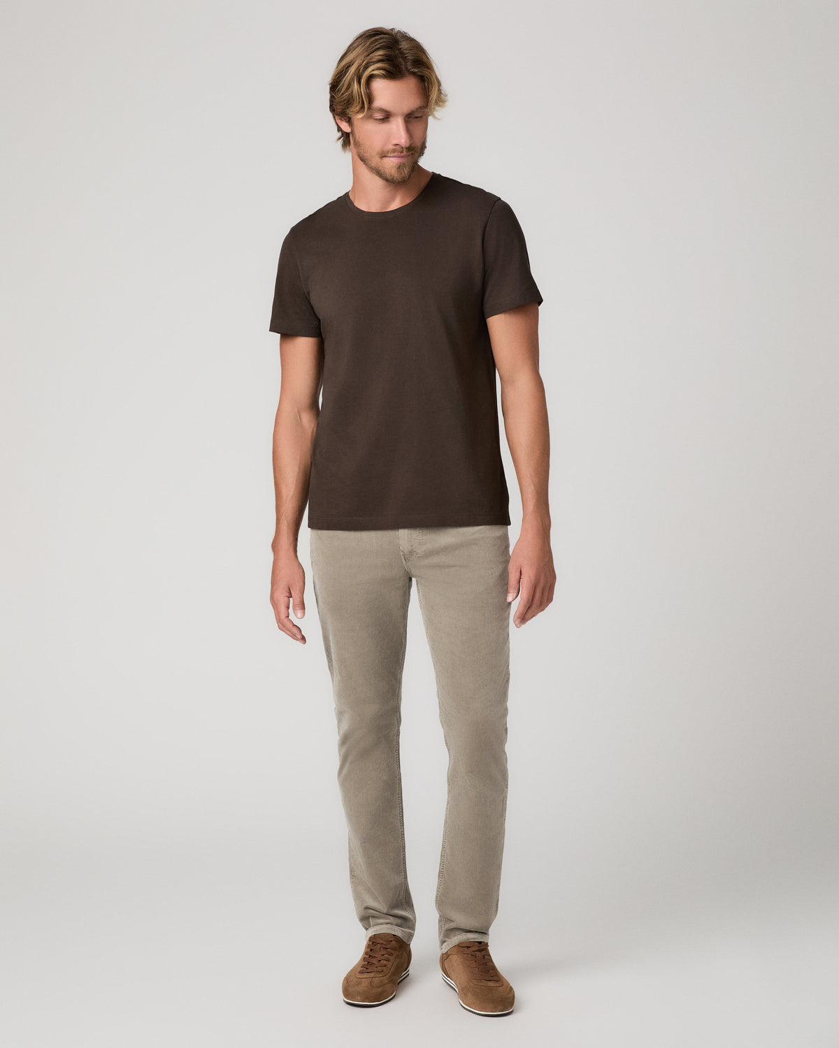 A man modeling a dark brown short-sleeve crewneck t-shirt, paired with beige trousers and brown suede shoes, standing against a light gray studio background.