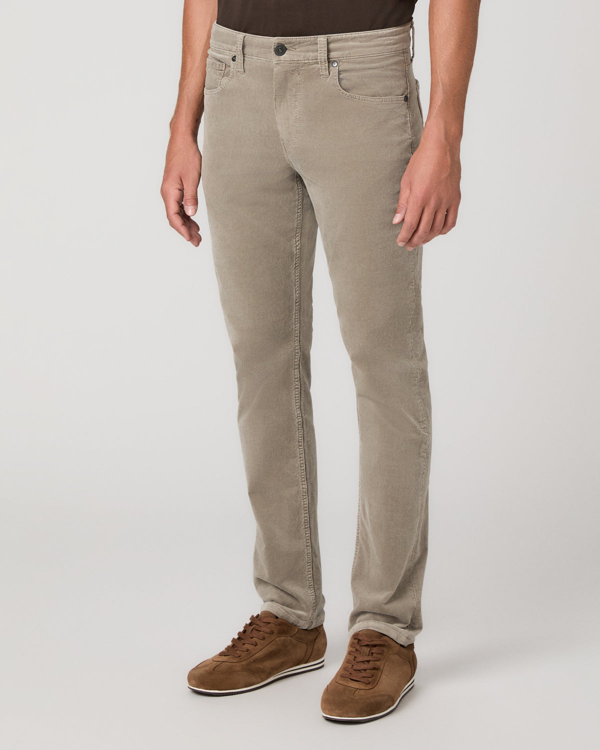 A man modeling a dark brown short-sleeve crewneck t-shirt, paired with beige trousers and brown suede shoes, standing against a light gray studio background.