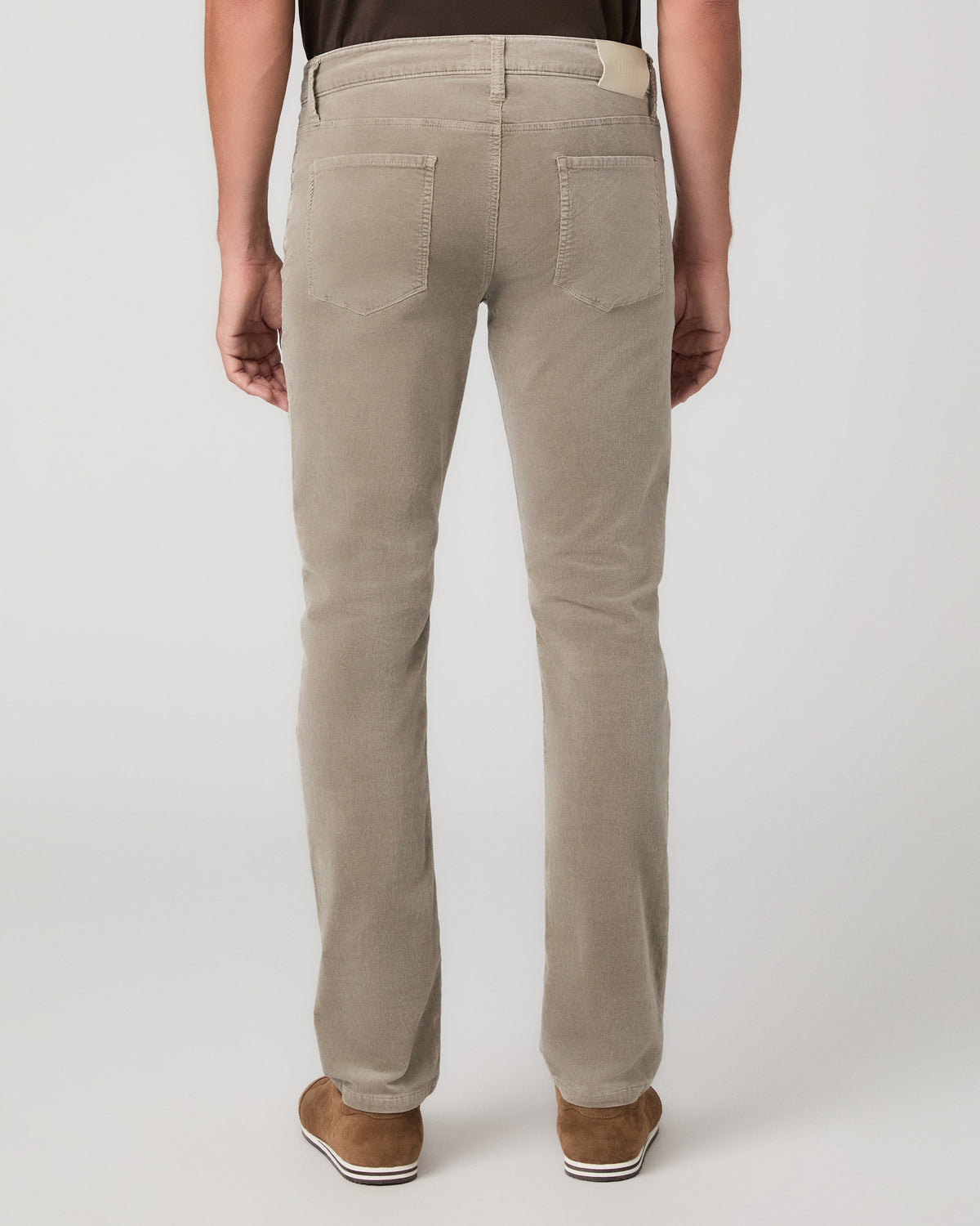 A man modeling a dark brown short-sleeve crewneck t-shirt, paired with beige trousers and brown suede shoes, standing against a light gray studio background.