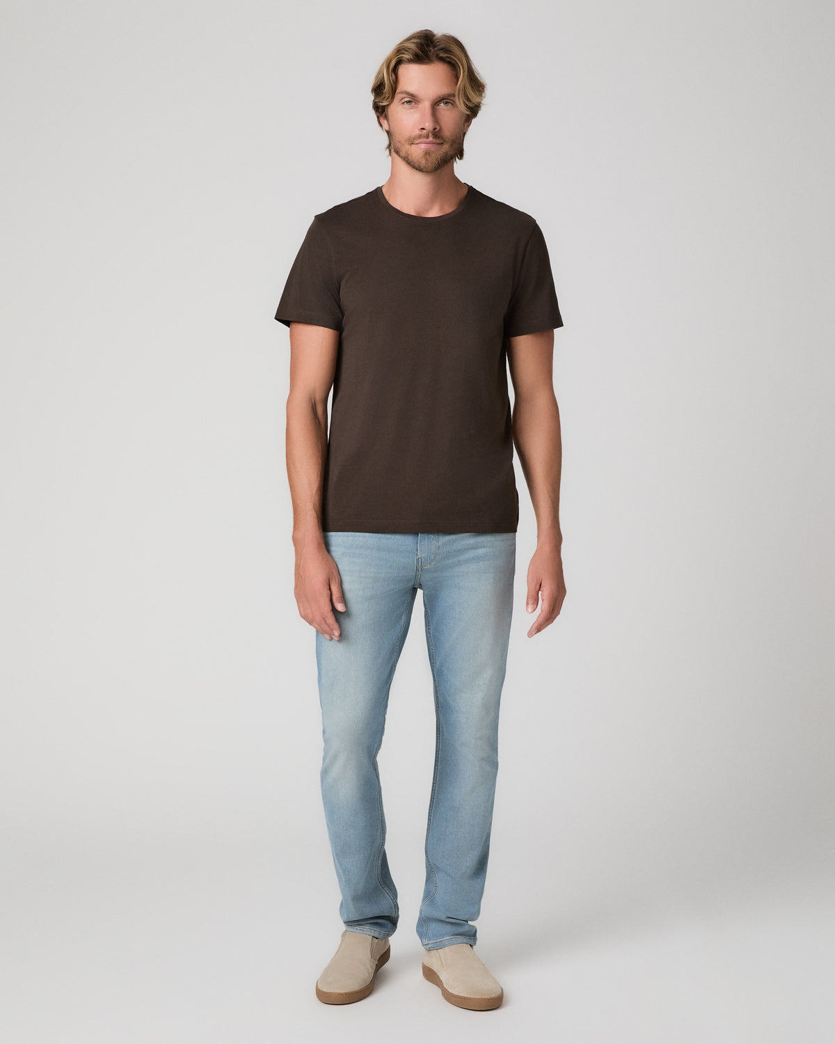 A man modeling a dark brown short-sleeve crewneck t-shirt, paired with light blue jeans and beige slip-on shoes, against a plain light gray studio background.