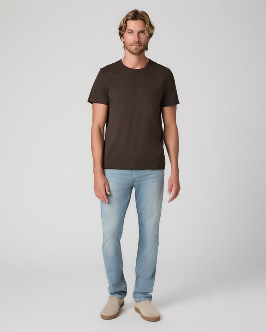 A man modeling a dark brown short-sleeve crewneck t-shirt, paired with light blue jeans and beige slip-on shoes, against a plain light gray studio background.
