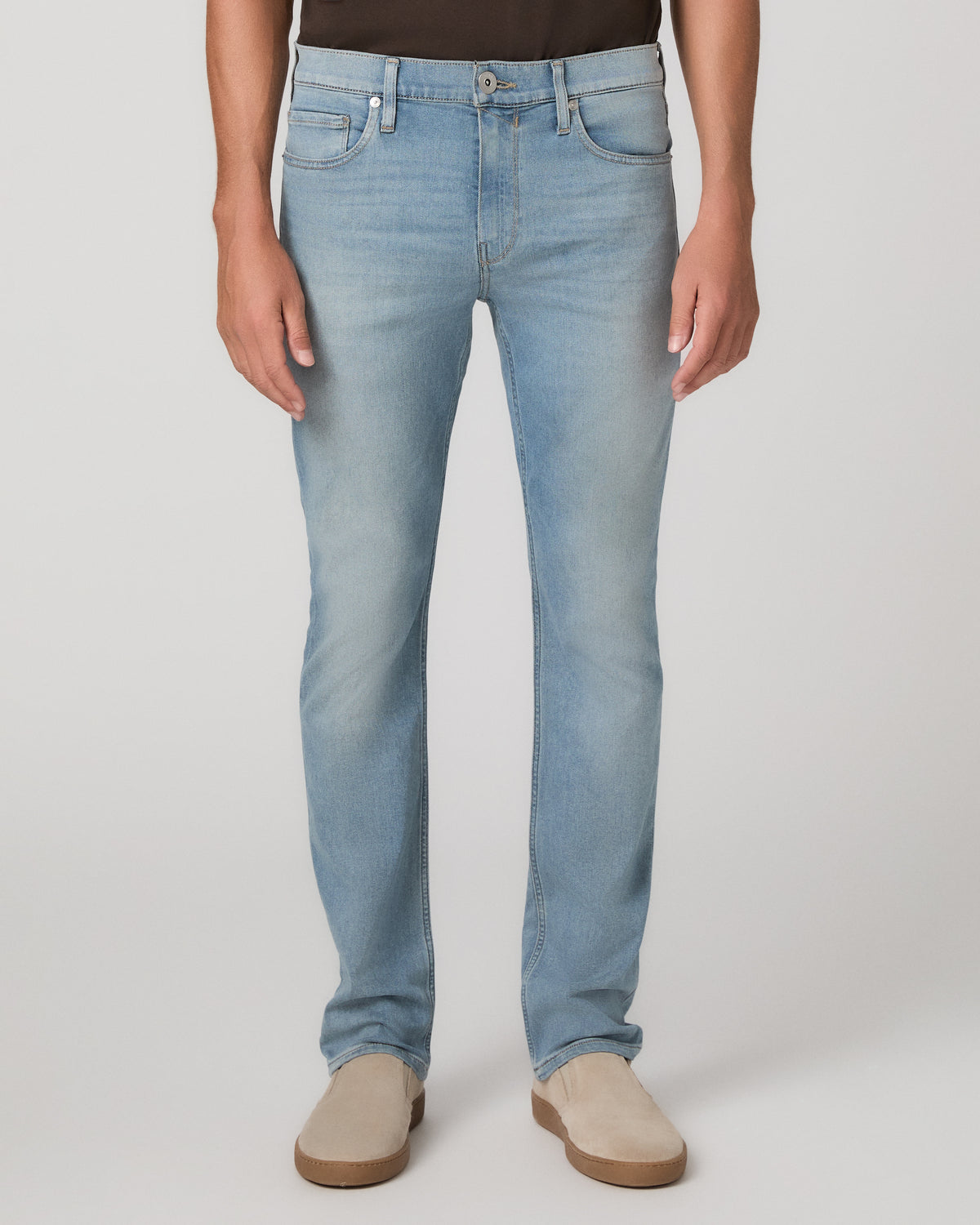 A man modeling a dark brown short-sleeve crewneck t-shirt, paired with light blue jeans and beige slip-on shoes, against a plain light gray studio background.