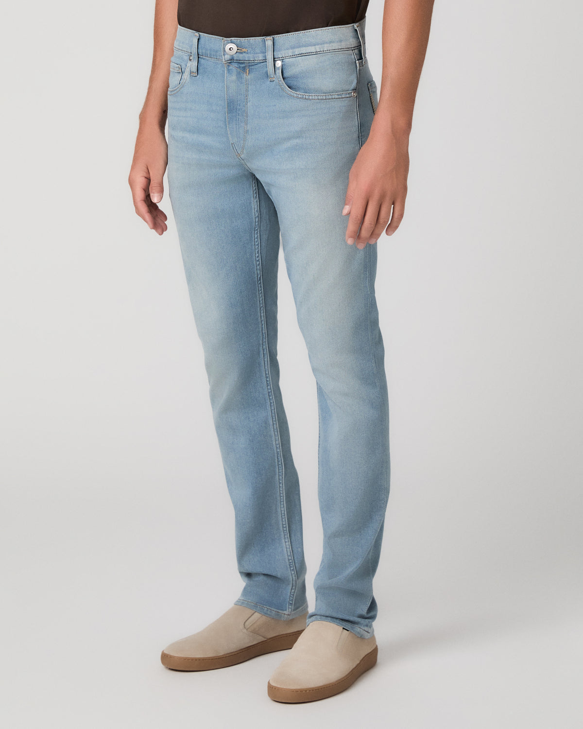 A man modeling a dark brown short-sleeve crewneck t-shirt, paired with light blue jeans and beige slip-on shoes, against a plain light gray studio background.