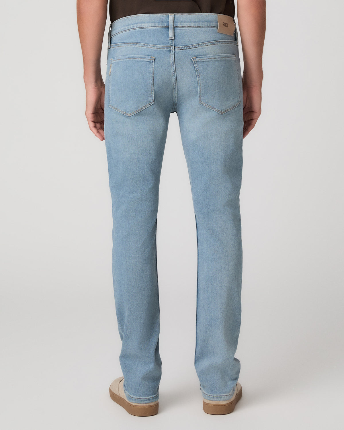 A man modeling a dark brown short-sleeve crewneck t-shirt, paired with light blue jeans and beige slip-on shoes, against a plain light gray studio background.