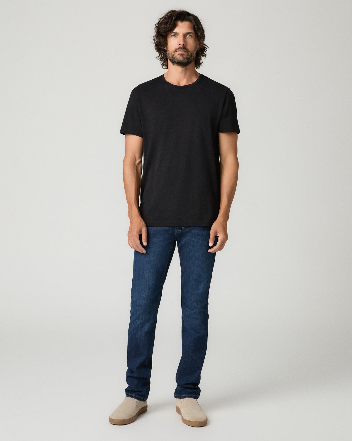 Full-length front view of a man with wavy dark hair wearing a black crew-neck t-shirt tucked into deep blue slim-fit jeans with subtle fading, paired with tan suede slip-on shoes.
