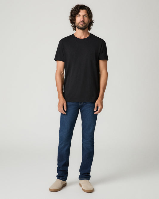 Full-length front view of a man with wavy dark hair wearing a black crew-neck t-shirt tucked into deep blue slim-fit jeans with subtle fading, paired with tan suede slip-on shoes.