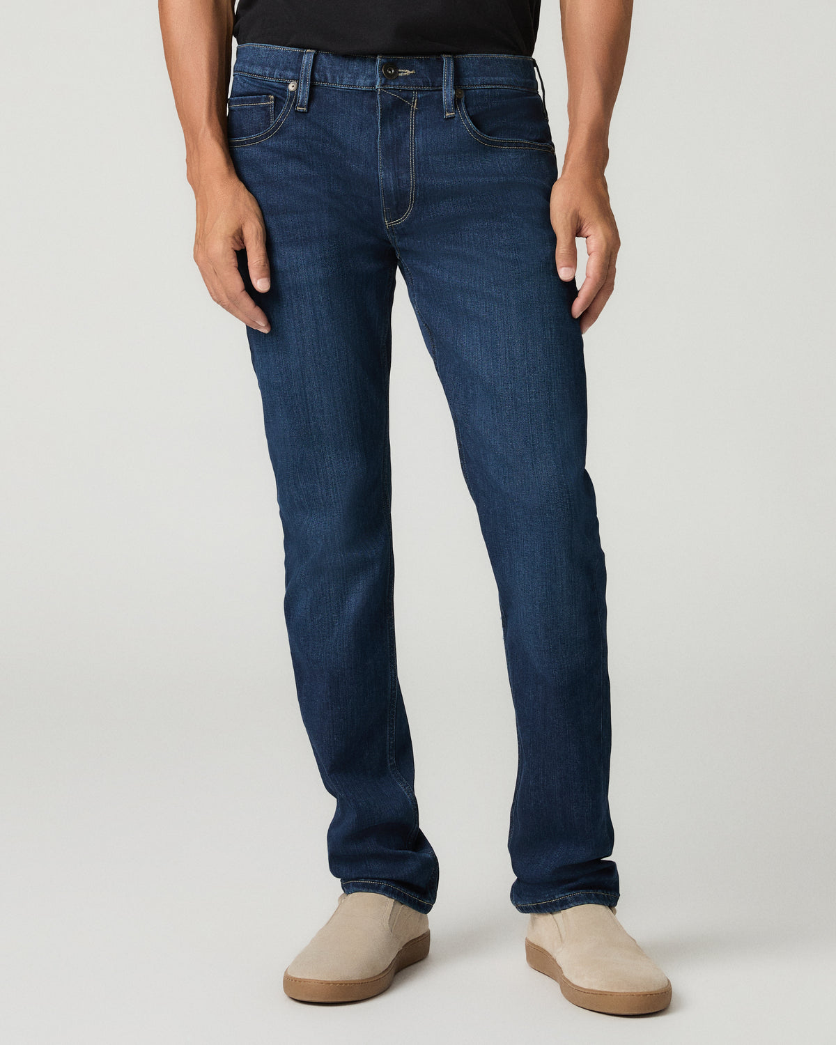Close-up front view of a man wearing deep blue slim-fit jeans with a button fly and five-pocket styling, cropped black t-shirt above and tan suede slip-on shoes below.