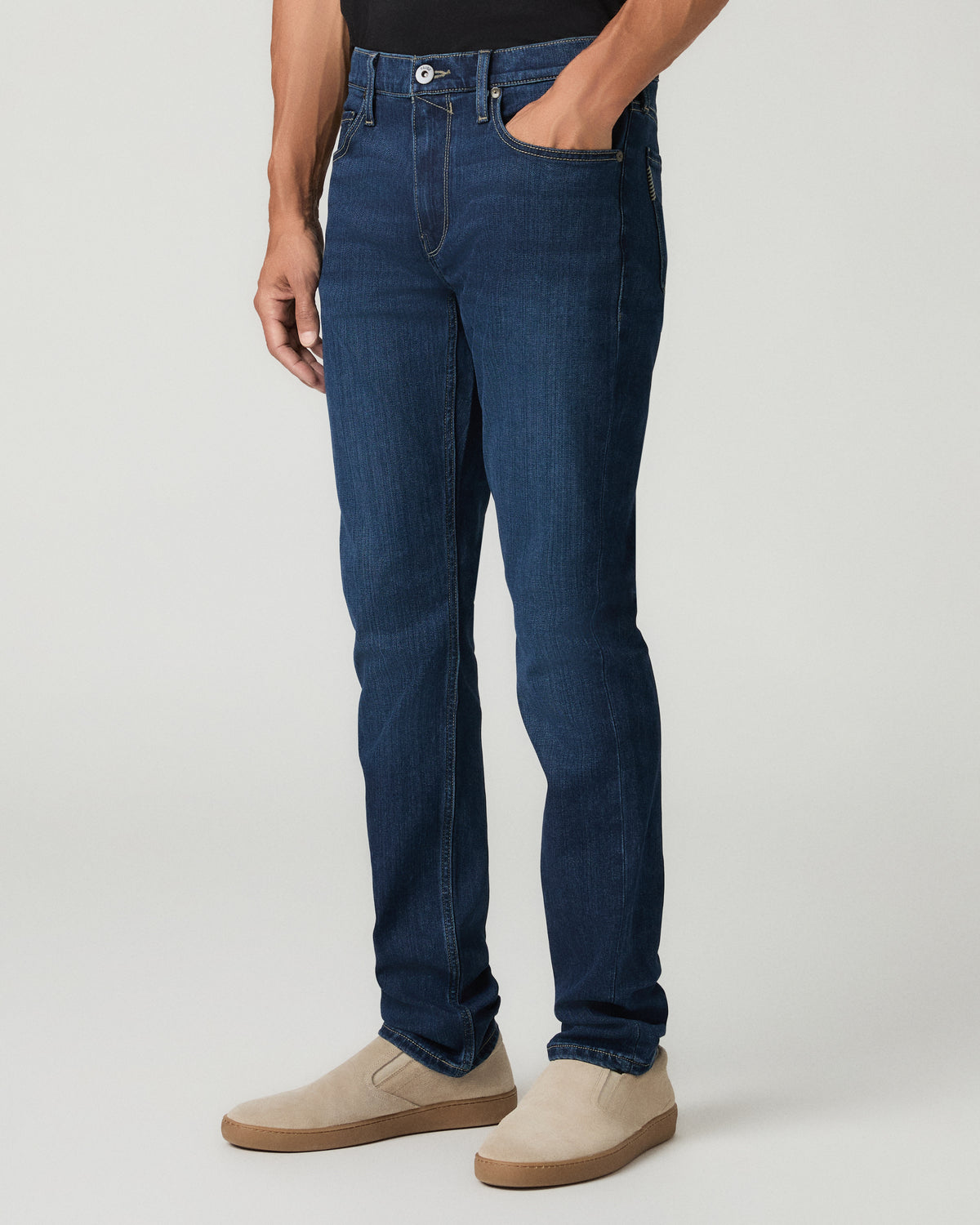Three-quarter angle view from the waist down of a man wearing deep blue slim-fit jeans with one hand tucked into the front pocket, paired with tan suede slip-on shoes.
