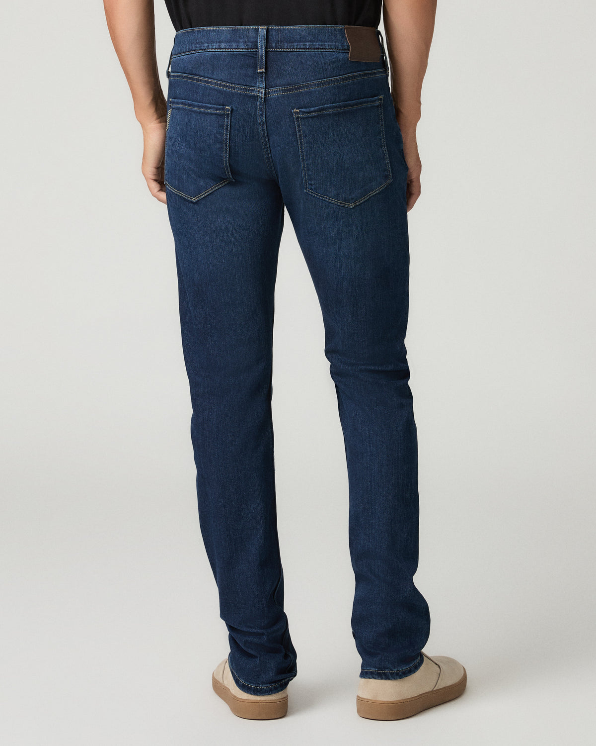 Full-length back view of a man wearing deep blue slim-fit jeans with a leather brand patch above the back pockets, black t-shirt, and tan suede slip-on shoes.