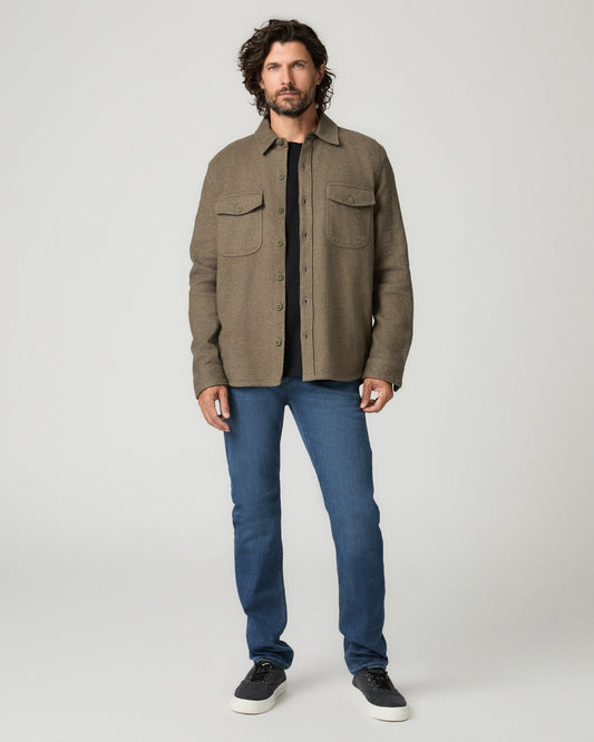 Full-length front view of a man with short dark hair wearing a heather gray ribbed knit sweater over a white collared shirt, tucked into medium blue slim-fit jeans, paired with black leather dress shoes.