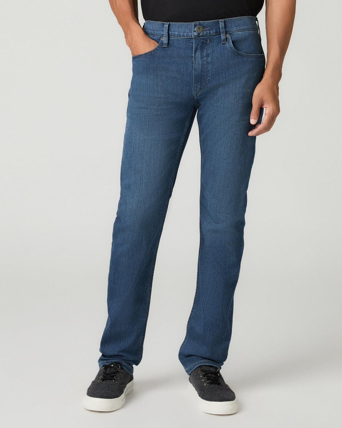 Close-up front view of a man wearing medium blue slim-fit jeans with subtle whisker fading at the thighs, cream sweater above and black leather shoes below.
