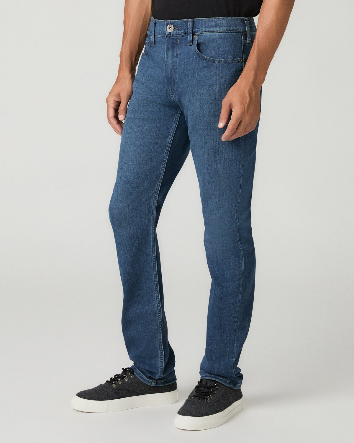 Three-quarter angle view of a man wearing medium blue slim-fit jeans and black leather dress shoes, standing with a relaxed stance.