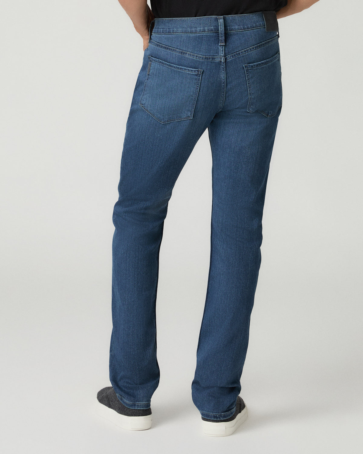 Full-length back view of a man wearing medium blue slim-fit jeans with a leather brand patch above the back pockets and black leather dress shoes.