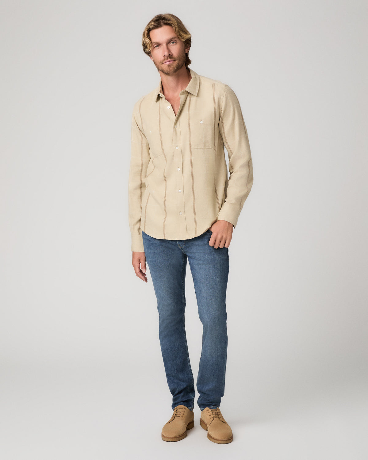 A man modeling a light beige long-sleeve button-up shirt, paired with blue jeans and tan boots, against a plain light gray studio background.