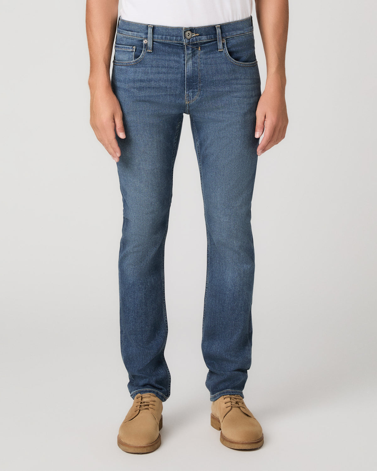 A man modeling a white tee paired with blue jeans and tan boots, against a plain light gray studio background.