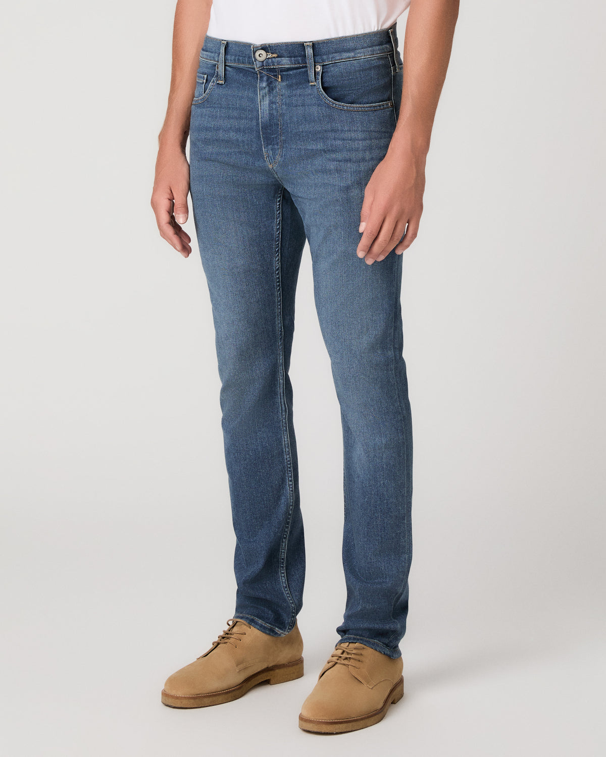 A man modeling a white tee paired with blue jeans and tan boots, against a plain light gray studio background.