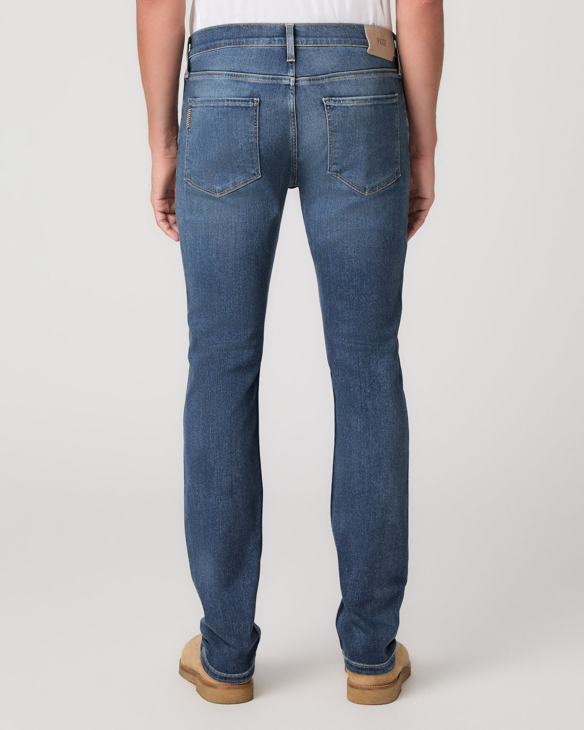 A man modeling a white tee paired with blue jeans and tan boots, against a plain light gray studio background.
