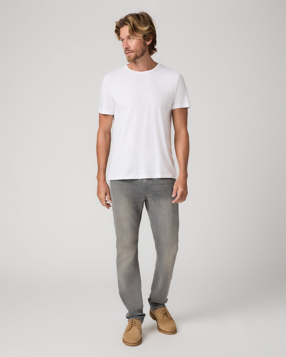 A man modeling a white short-sleeve crewneck t-shirt, paired with gray slim-fit jeans and tan suede shoes, standing against a light gray studio background.