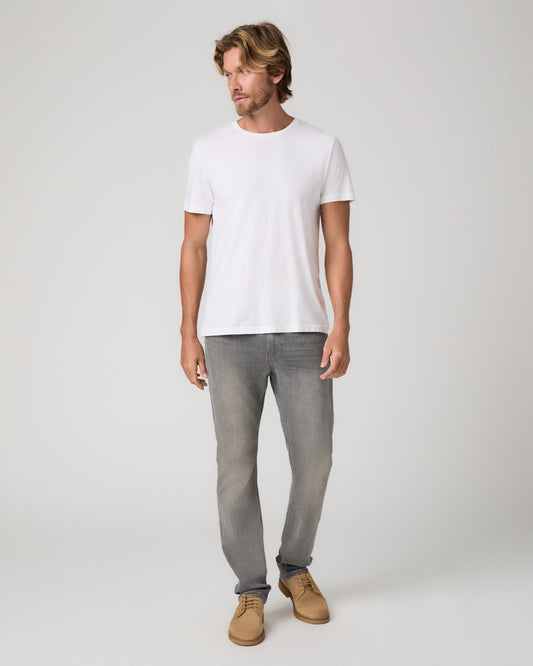 A man modeling a white short-sleeve crewneck t-shirt, paired with gray slim-fit jeans and tan suede shoes, standing against a light gray studio background.