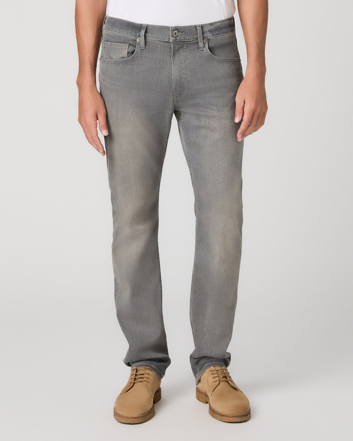 A man modeling a white short-sleeve crewneck t-shirt, paired with gray slim-fit jeans and tan suede shoes, standing against a light gray studio background.