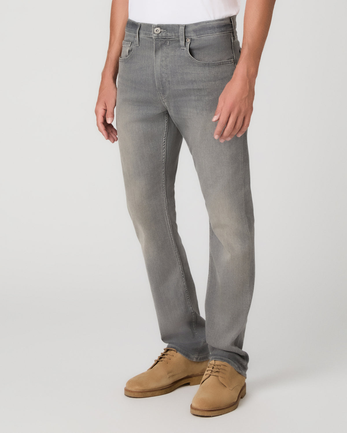 A man modeling a white short-sleeve crewneck t-shirt, paired with gray slim-fit jeans and tan suede shoes, standing against a light gray studio background.