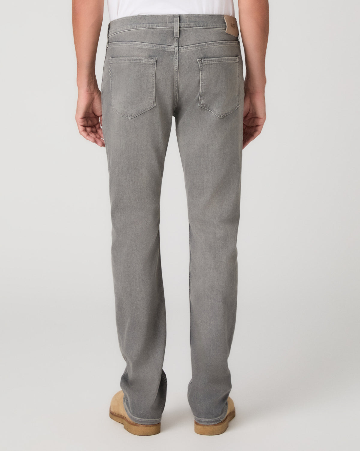A man modeling a white short-sleeve crewneck t-shirt, paired with gray slim-fit jeans and tan suede shoes, standing against a light gray studio background.