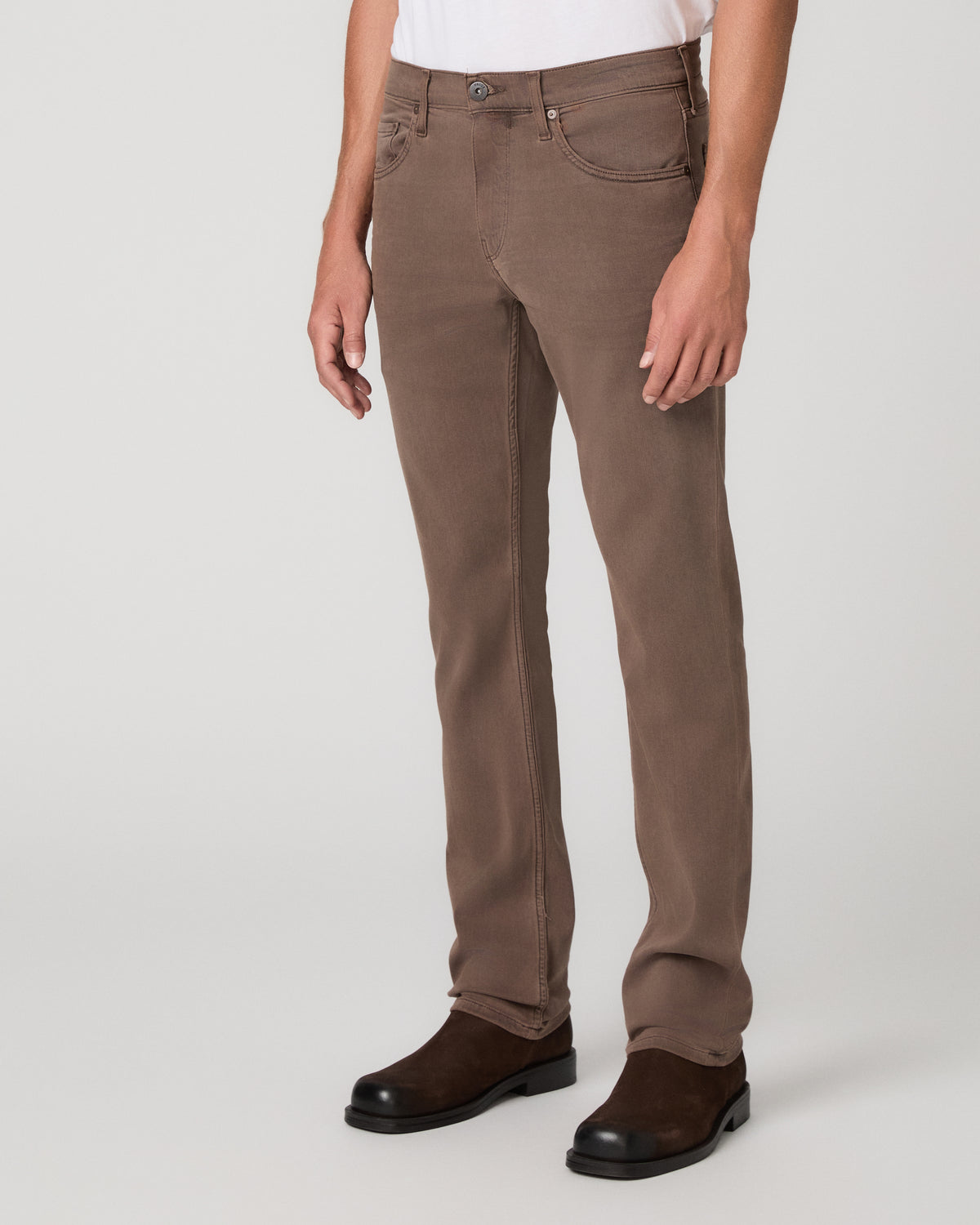 A man modeling a white tee and matching brown trousers paired with brown shoes against a light gray studio background.