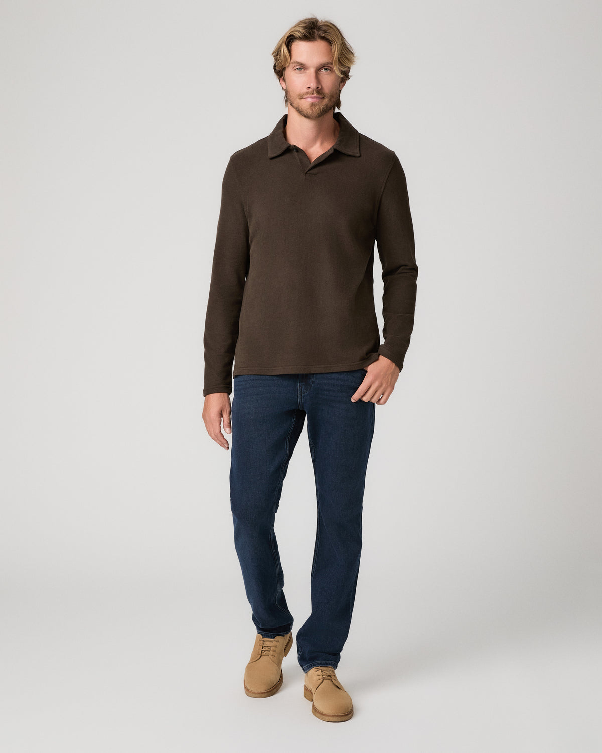 A man modeling a dark brown long-sleeve knit polo shirt, paired with dark blue jeans and tan suede boots, against a light gray studio background.