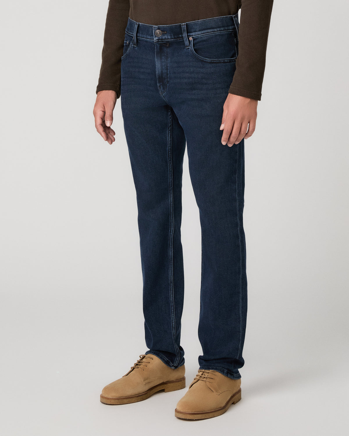A man modeling a dark brown long-sleeve knit polo shirt, paired with dark blue jeans and tan suede boots, against a light gray studio background.