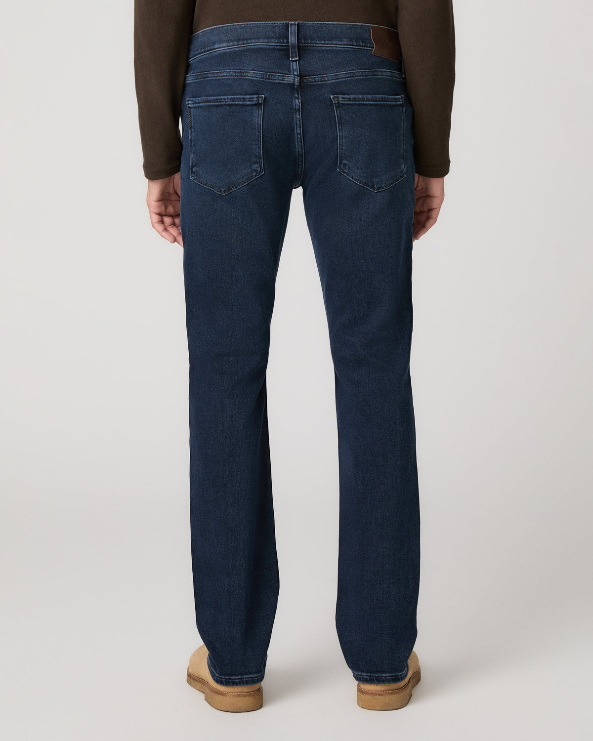 A man modeling a dark brown long-sleeve knit polo shirt, paired with dark blue jeans and tan suede boots, against a light gray studio background.