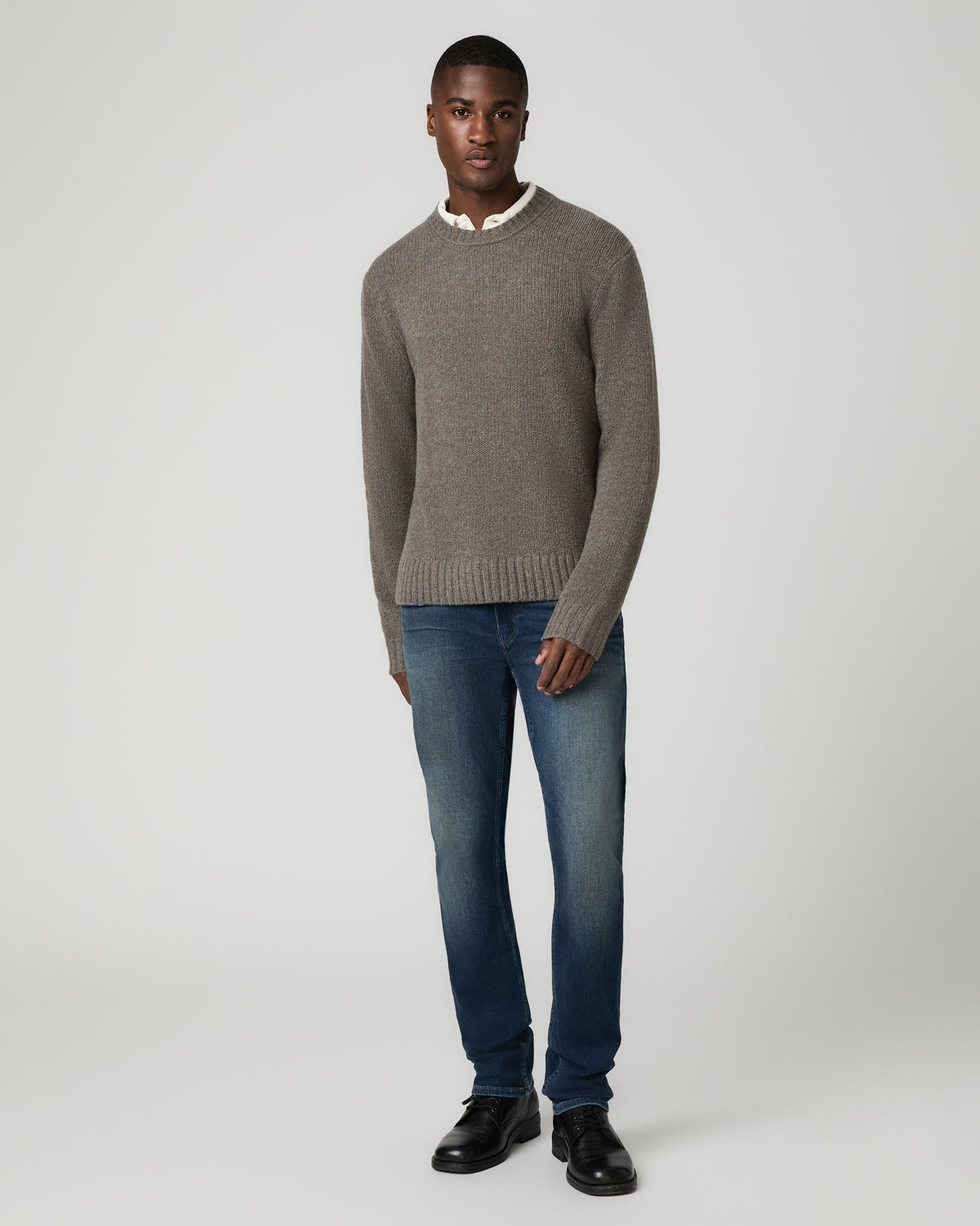 Full-length front view of a man wearing a heather grey chunky knit crew-neck sweater, paired with medium-dark blue wash Minden slim straight jeans and black leather shoes.