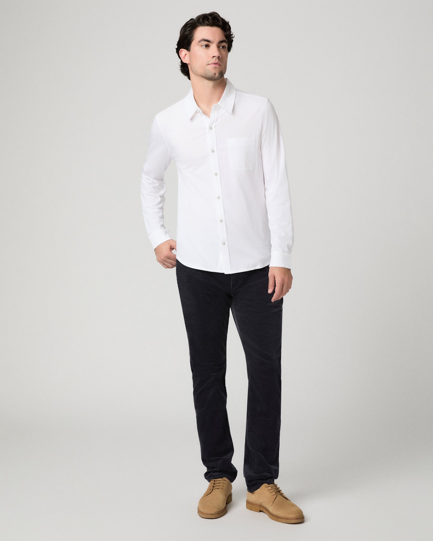 A man modeling a white long-sleeve button-down shirt with a chest pocket, paired with dark trousers and tan suede shoes, against a light gray studio background.