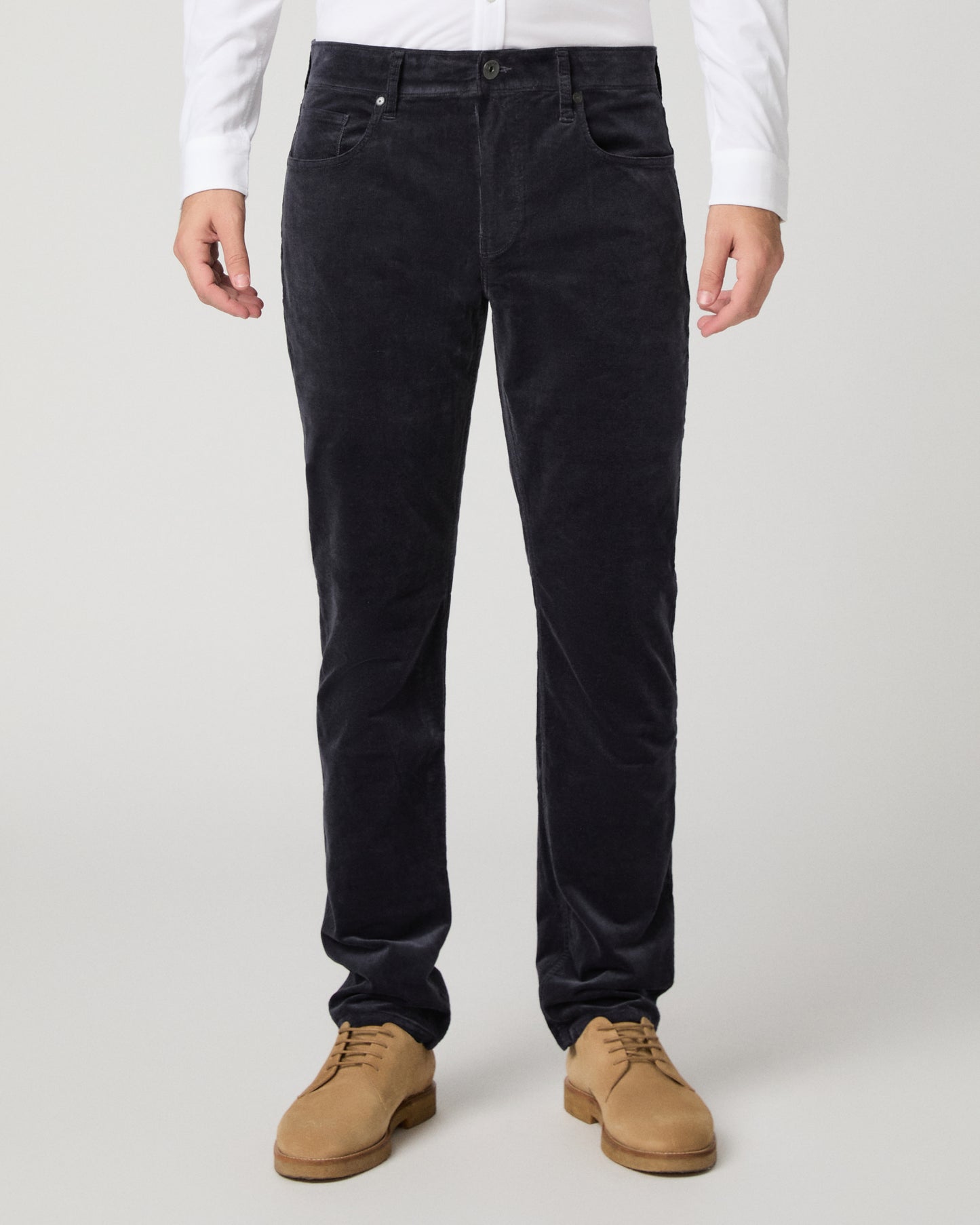 A person wearing slim-fit black corduroy trousers, styled with a white long-sleeve shirt and tan suede boots, photographed against a light gray studio background.