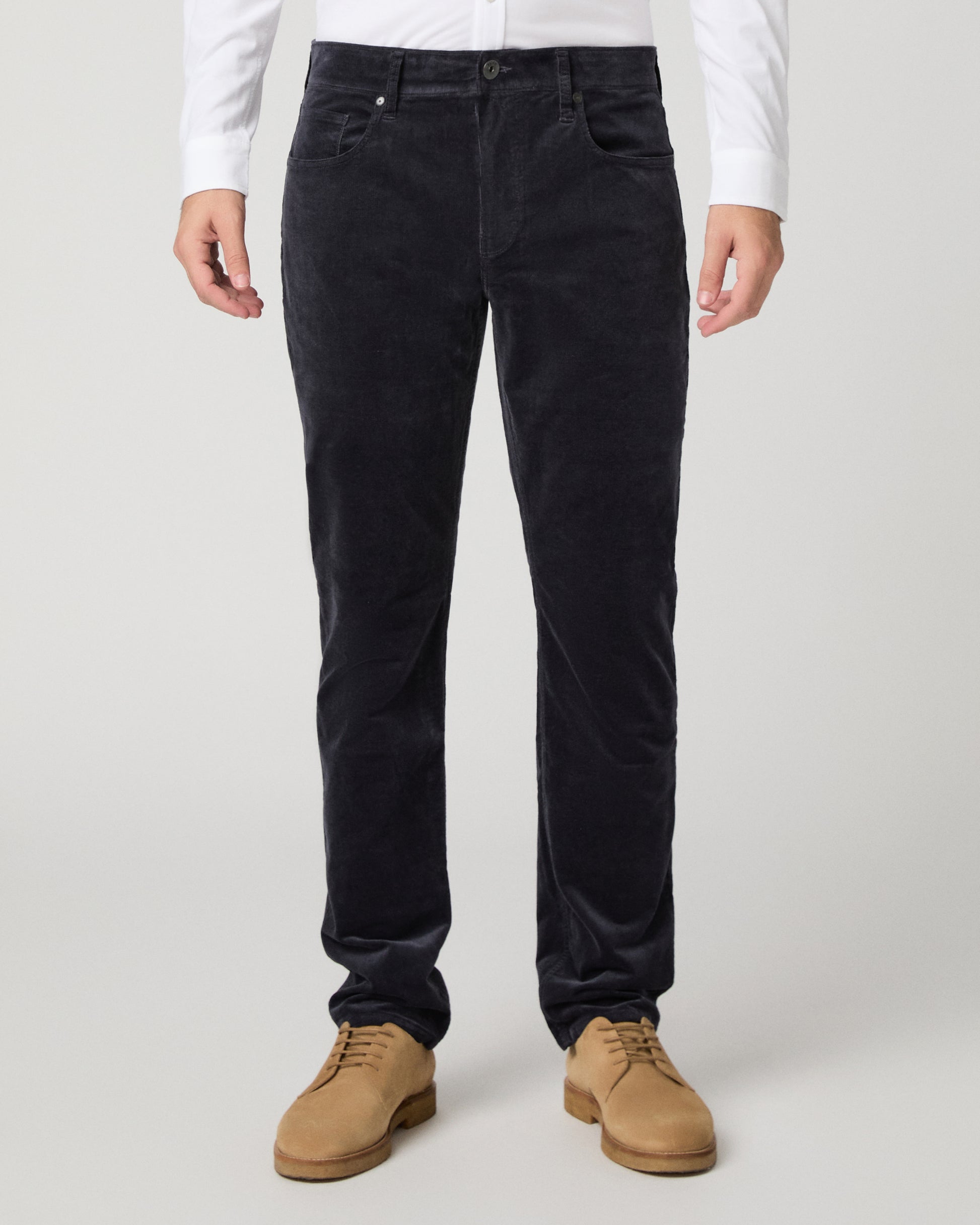 A person wearing slim-fit black corduroy trousers, styled with a white long-sleeve shirt and tan suede boots, photographed against a light gray studio background.