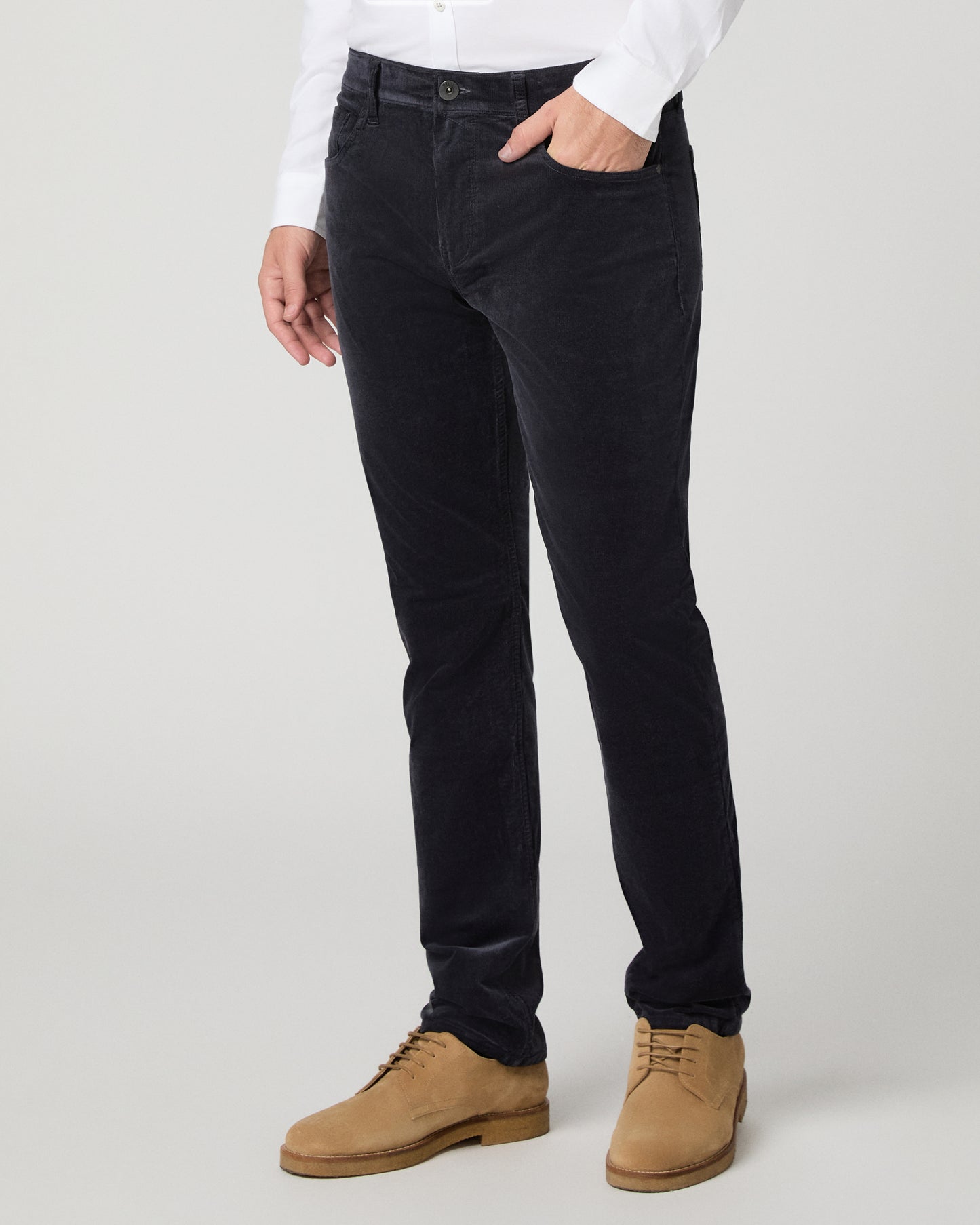 A person wearing slim-fit black corduroy trousers, styled with a white long-sleeve shirt and tan suede lace-up shoes, photographed against a light gray studio background.