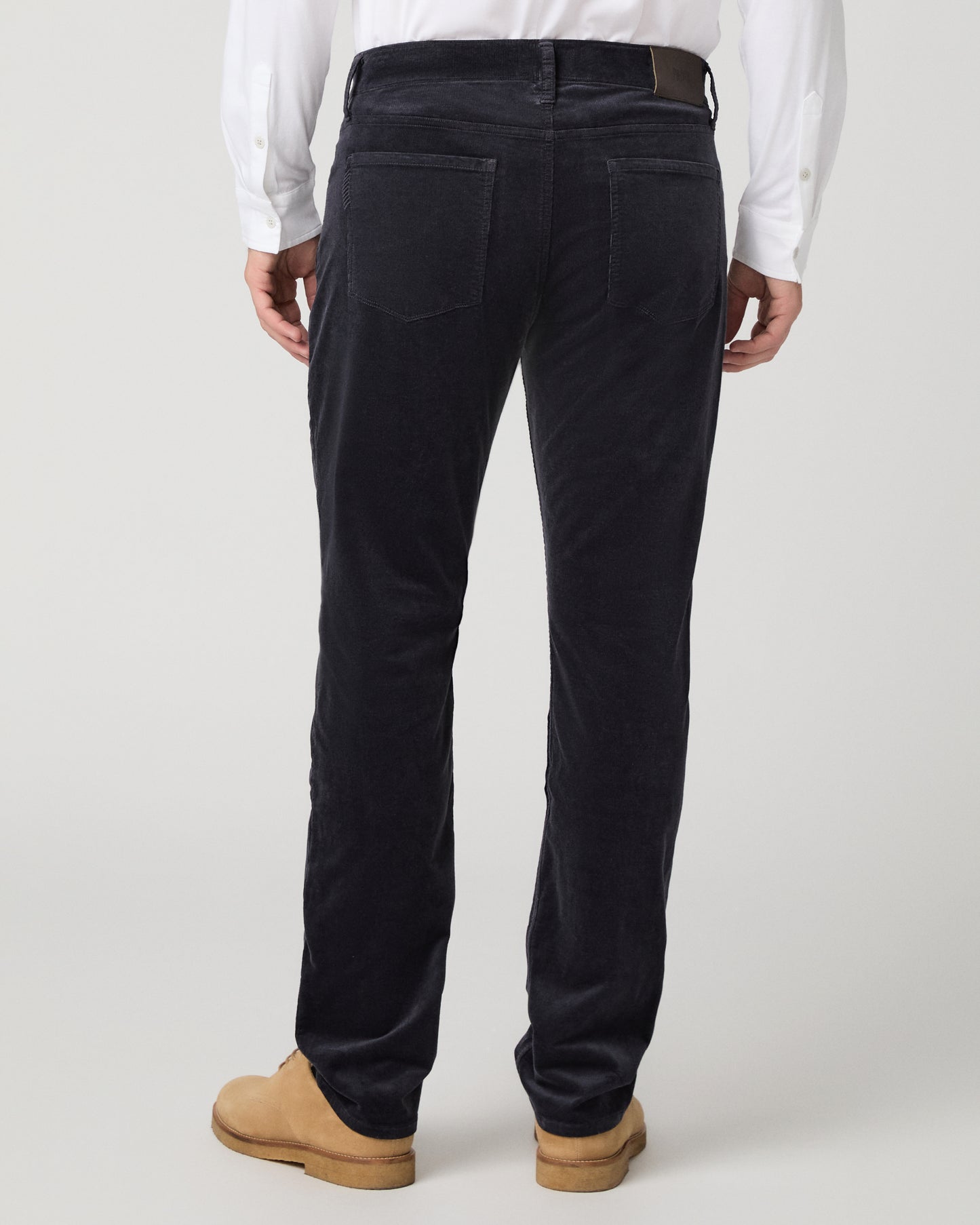 Rear view of a person wearing dark navy corduroy trousers, a white long-sleeve shirt, and tan suede boots against a light gray studio background.