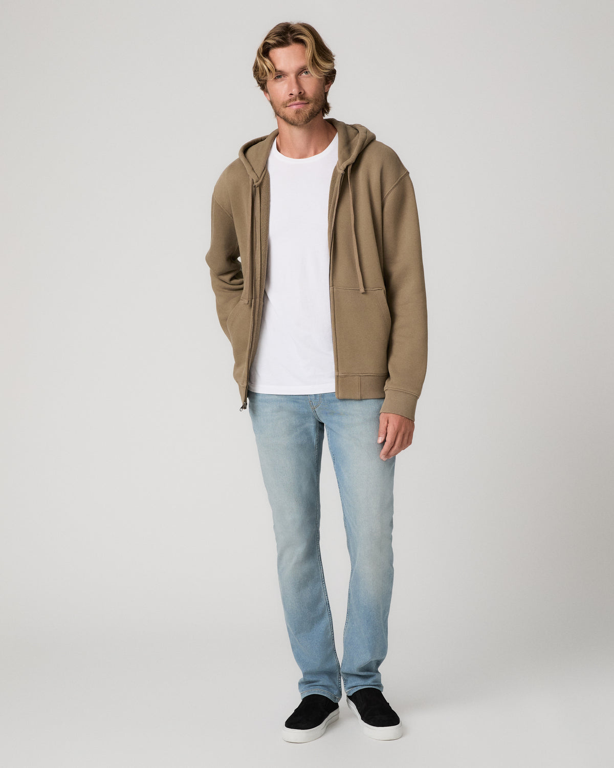 A man modeling a brown long sleeve sweater and white tee paired with light blue jeans and black sneakers, standing against a light gray studio background.