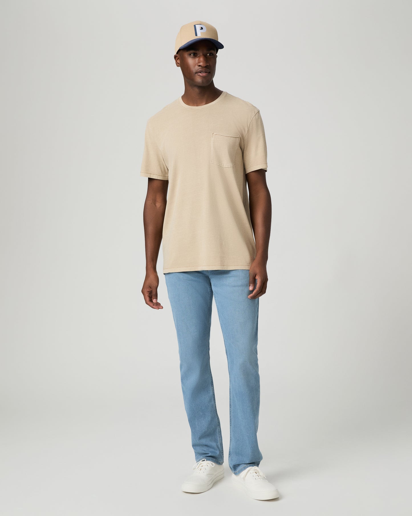 Man wearing a beige t-shirt, light blue jeans, and a beige cap with a logo on a white background