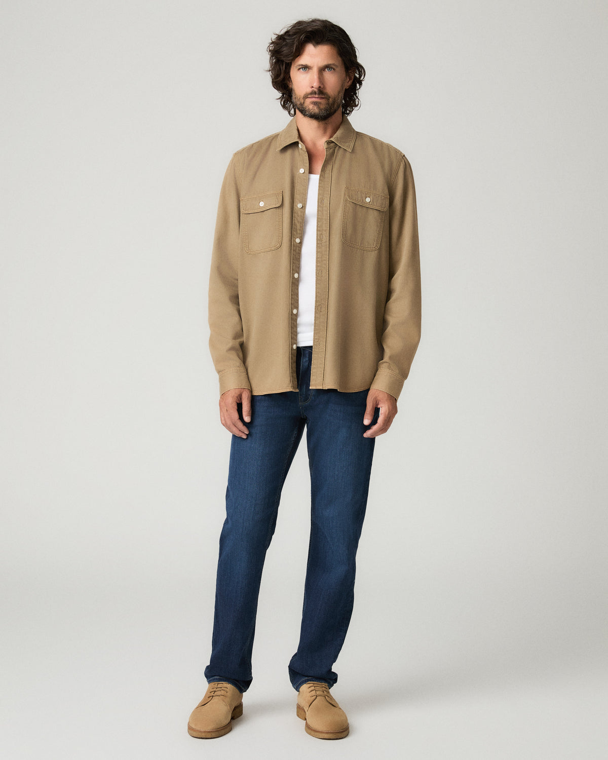 Full-length front view of a man with dark curly hair wearing an open tan button-front overshirt with chest pockets over a white t-shirt, one hand in his pocket, paired with deep blue slim-straight jeans and tan suede shoes.