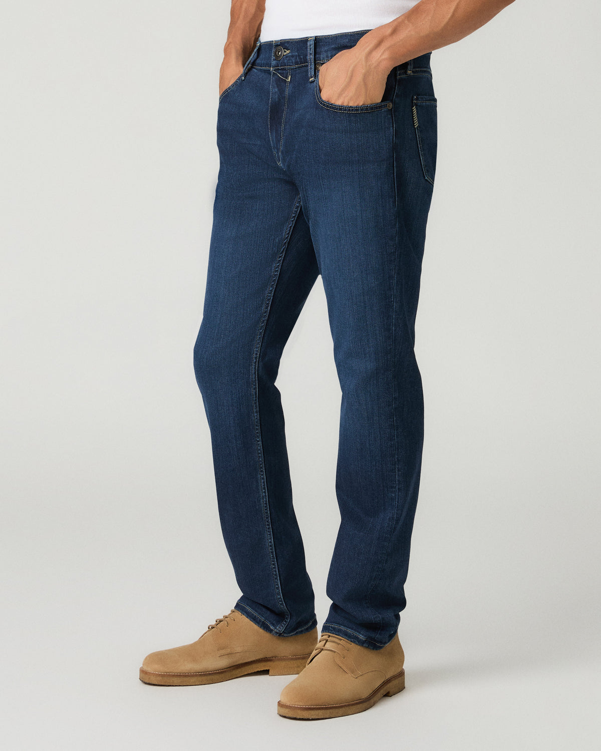 Three-quarter angle view of a man with one hand in his pocket wearing deep blue slim-straight jeans and tan suede shoes.